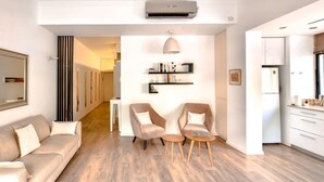A modern living room with a sofa, two armchairs, a coffee table, a bookshelf, and a kitchen area with cabinets and a window. - Rupin Nacre in Tel Aviv-yafo (Tel Aviv)