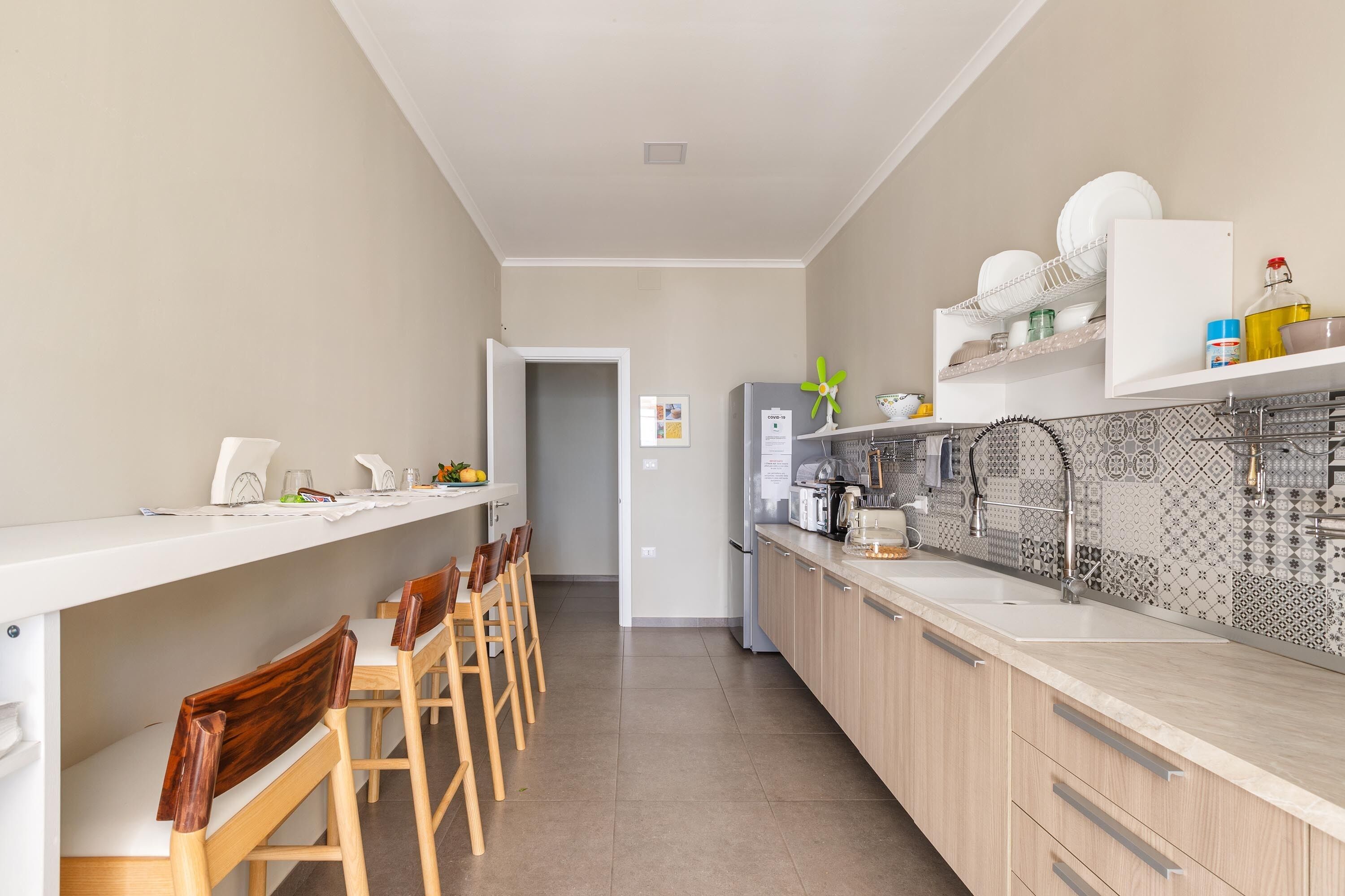 A modern kitchen with a long countertop, wooden chairs, a refrigerator, and patterned backsplash.