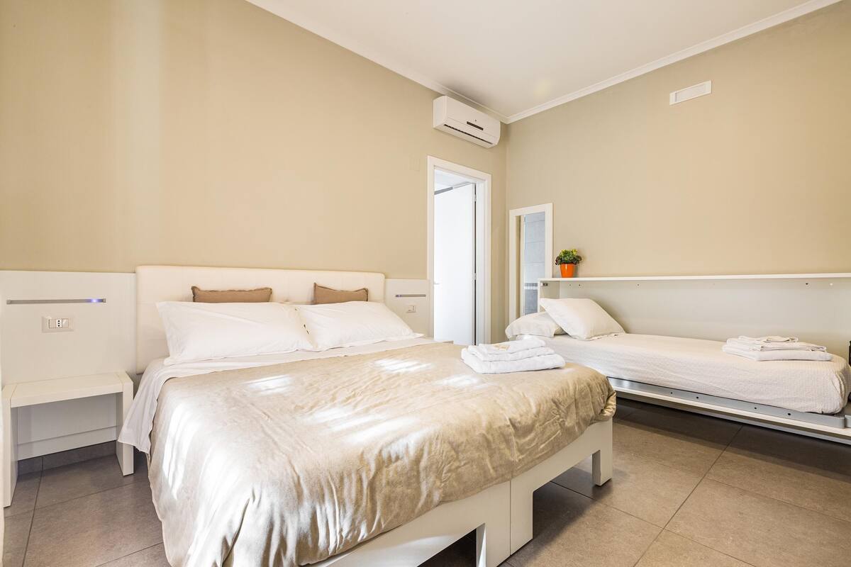 A hotel room with a large bed, a smaller bed, a bedside table, a nightstand, a mirror, and an air conditioning unit.