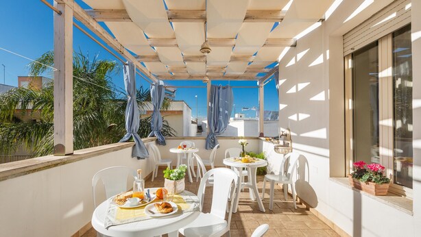 A sunlit outdoor dining area with white tables and chairs, a wooden pergola, and a view of palm trees and buildings. - 2674 B&B Albatro - Camera Tripla (Taviano)