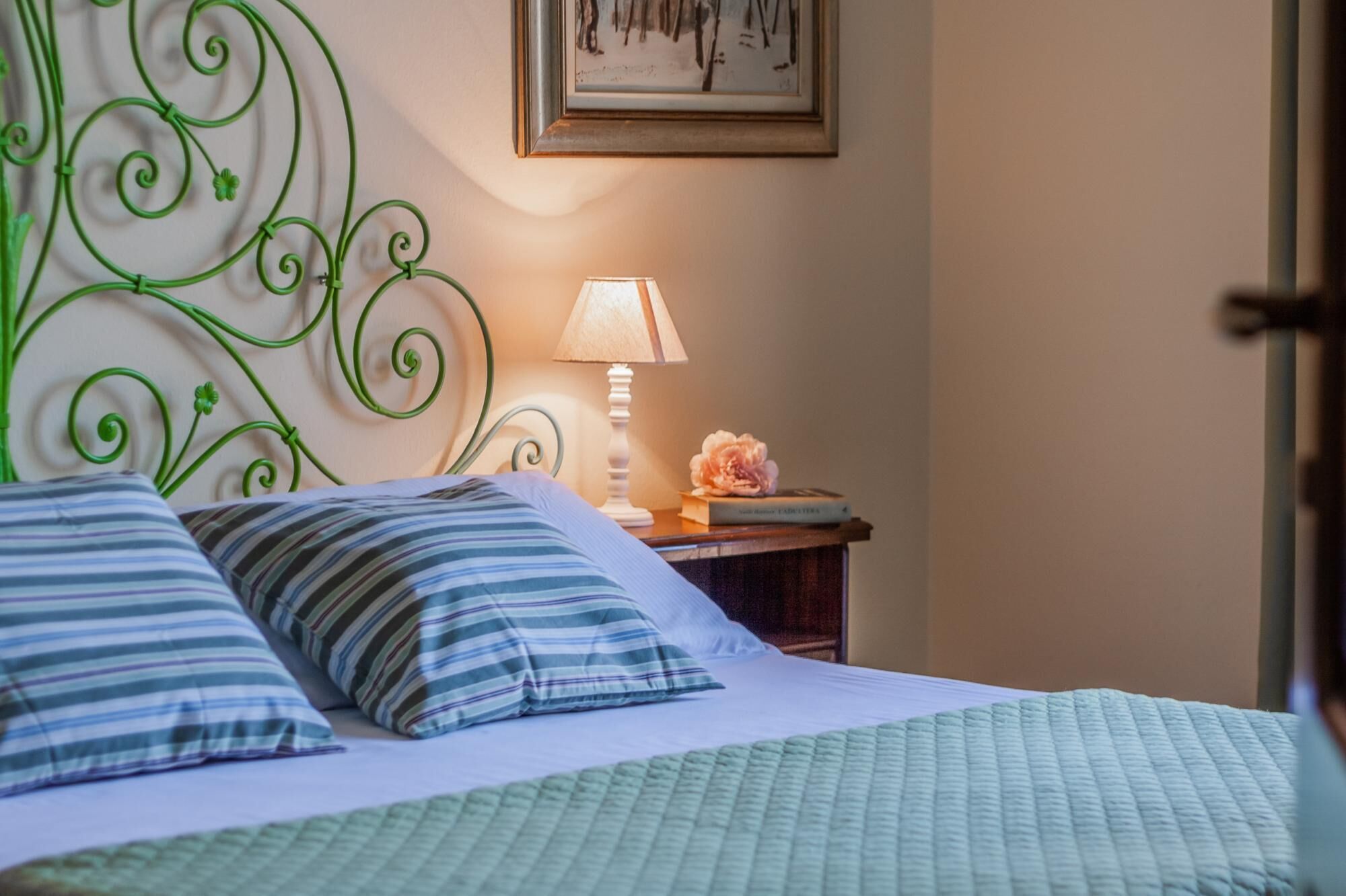 A neatly made bed with striped pillows and a green quilt, a bedside table with a lamp and a book, and a framed picture on the wall.