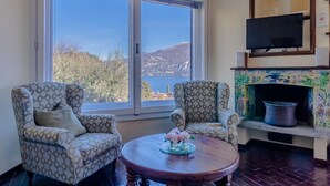 Una sala de estar con dos sillones estampados, una mesa de centro redonda de madera con un centro de mesa floral, una chimenea con detalles de azulejos y un ventanal grande que ofrece una vista panorámica de colinas y árboles.