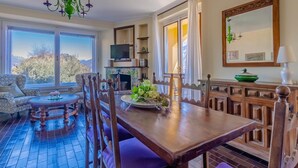 Un comedor con mesa de madera, sillas y una araña. Hay un televisor en un estante, una chimenea y una ventana con cortinas.