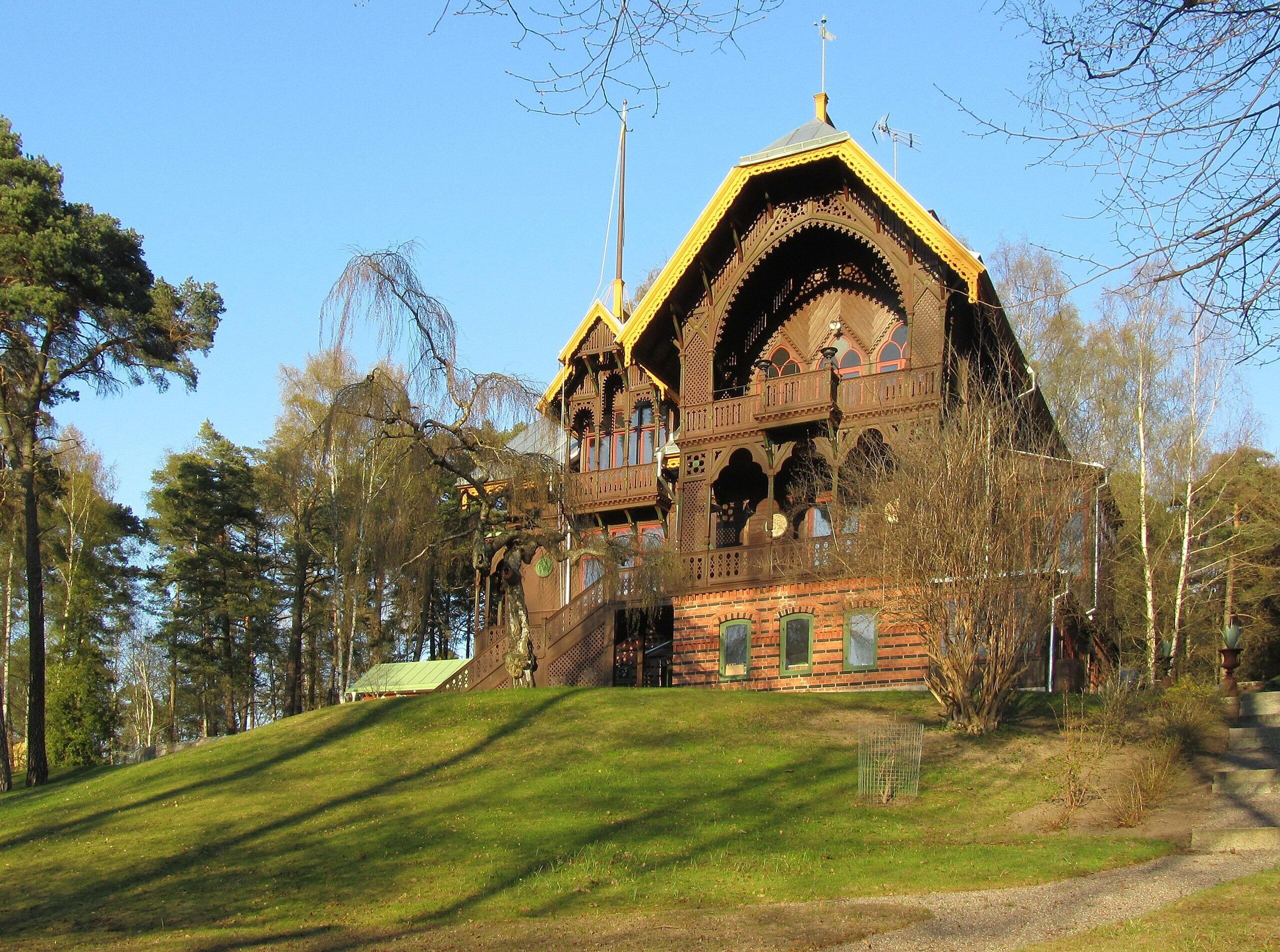 A large, ornate wooden building with multiple balconies and a steep roof, surrounded by trees and a grassy area.
