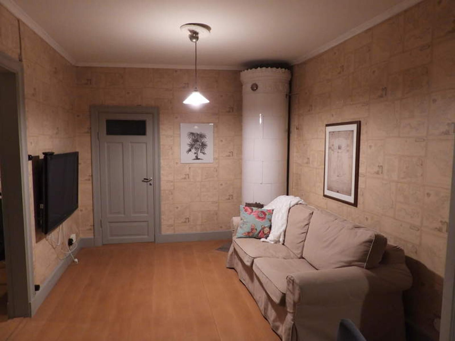 A room with a beige sofa, a flat-screen TV mounted on the wall, a door, and a framed picture.
