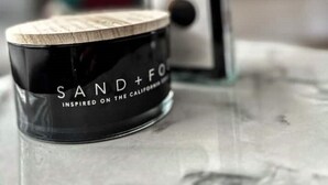 A black container with "SAND+FO" and "INSPIRED ON THE CALIFORNIA" text, a clear glass bottle, and a blurred background with a TV and shelves. - Comfy house in West Lothian - 10 min from the airport! (Broxburn)