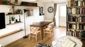 A cozy living room with a wooden dining table, two chairs, a bookshelf, a television, and a large window with curtains. - Comfy house in West Lothian - 10 min from the airport! (Broxburn)