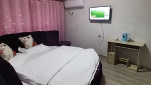 A bedroom with a bed, a TV mounted on the wall, a desk with a kettle, and a window with pink curtains.