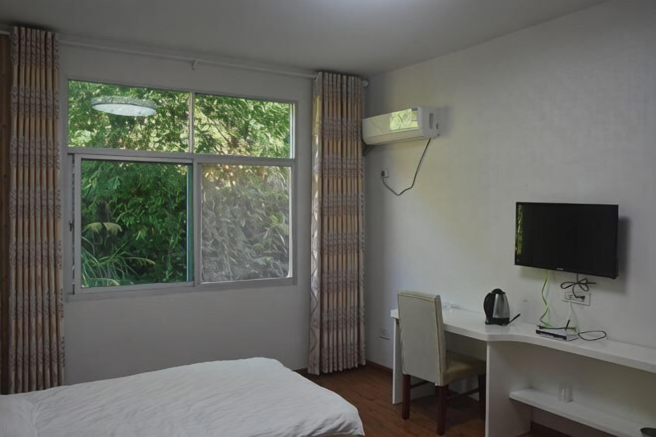 A bedroom with a bed, a desk with a chair, a TV, and a window with curtains.