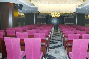 Une salle de conférence avec des rangées de chaises en bois et des coussins roses, un lustre et une scène à l’avant.