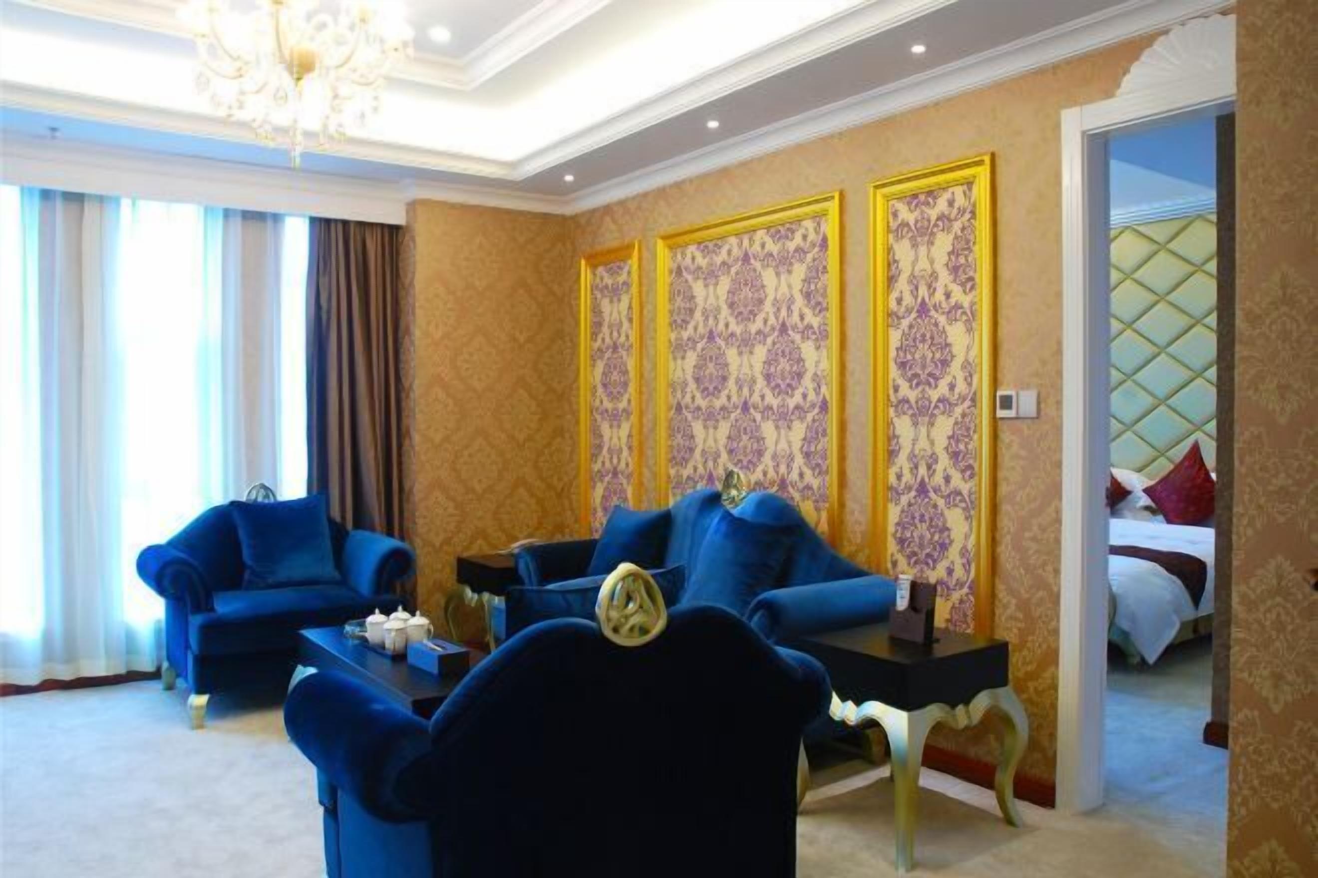 A hotel room with two blue armchairs, a small table, and a bed with a red pillow.