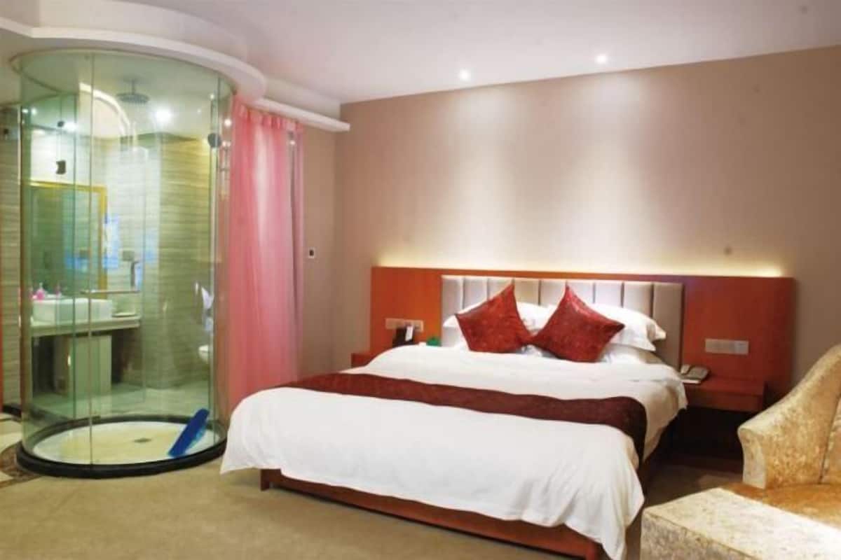 A hotel room with a glass-enclosed shower, a bed with white bedding and red pillows, and a beige armchair.