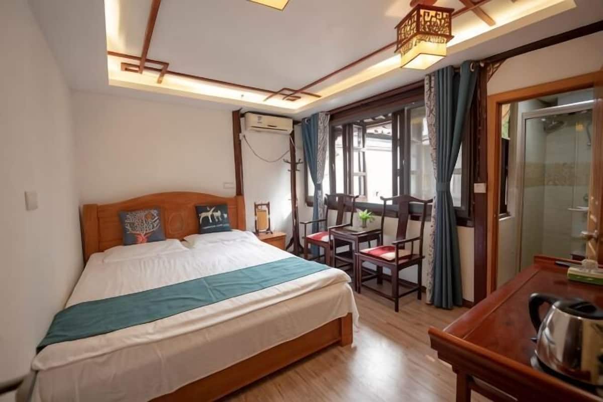 A bedroom with a bed, a desk, and a chair. There is a window with curtains, a ceiling fan, and a wall-mounted air conditioner.