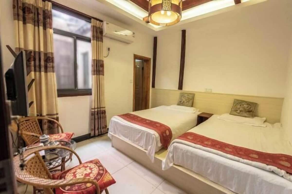 A hotel room with two beds, a wooden ceiling, a window with curtains, a table with chairs, and a wall-mounted air conditioner.