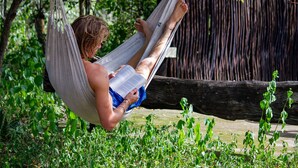 Una persona recostada en una hamaca leyendo un libro en un entorno natural, con follaje y una estructura de madera al fondo.