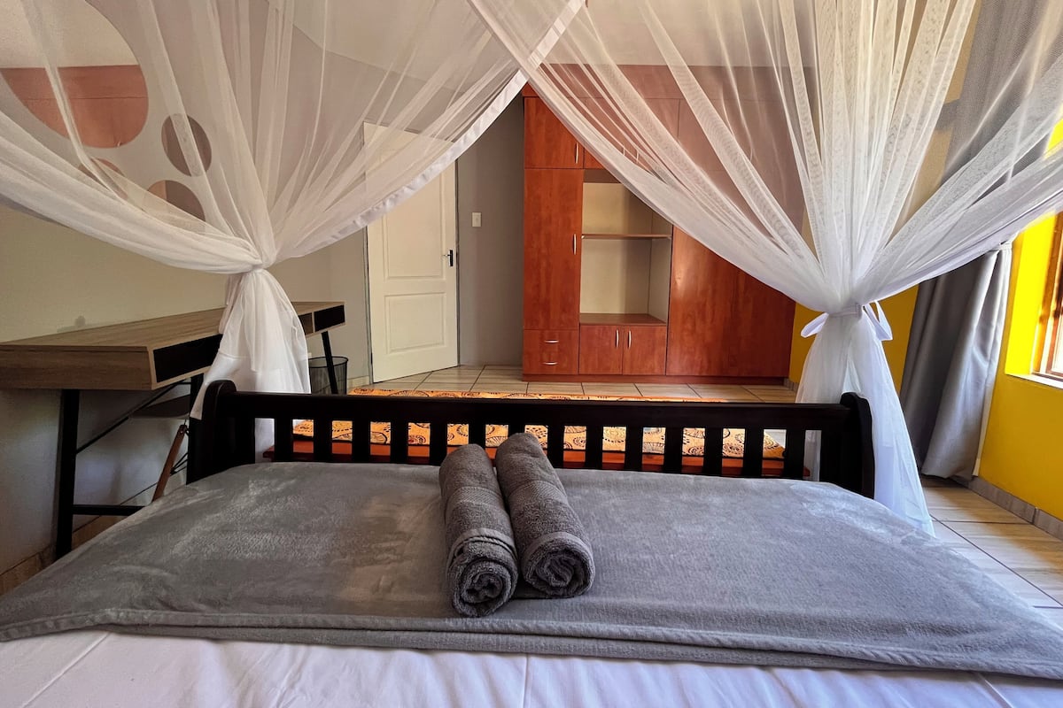 A bed with a grey blanket and two rolled towels, a wooden headboard, and a white canopy.