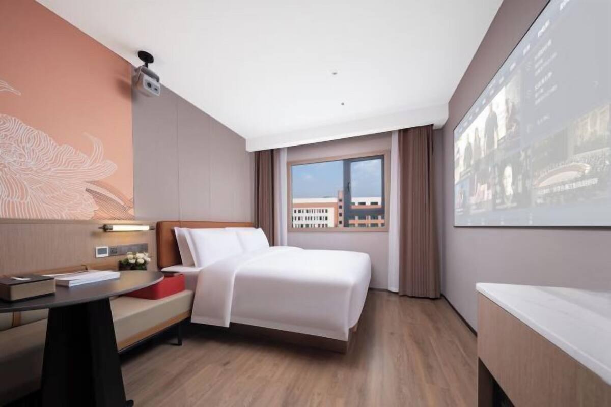 Une chambre d’hôtel équipée d’un lit, d’un bureau, d’une chaise, d’une fenêtre donnant sur des immeubles et d’un écran de projection fixé au mur.