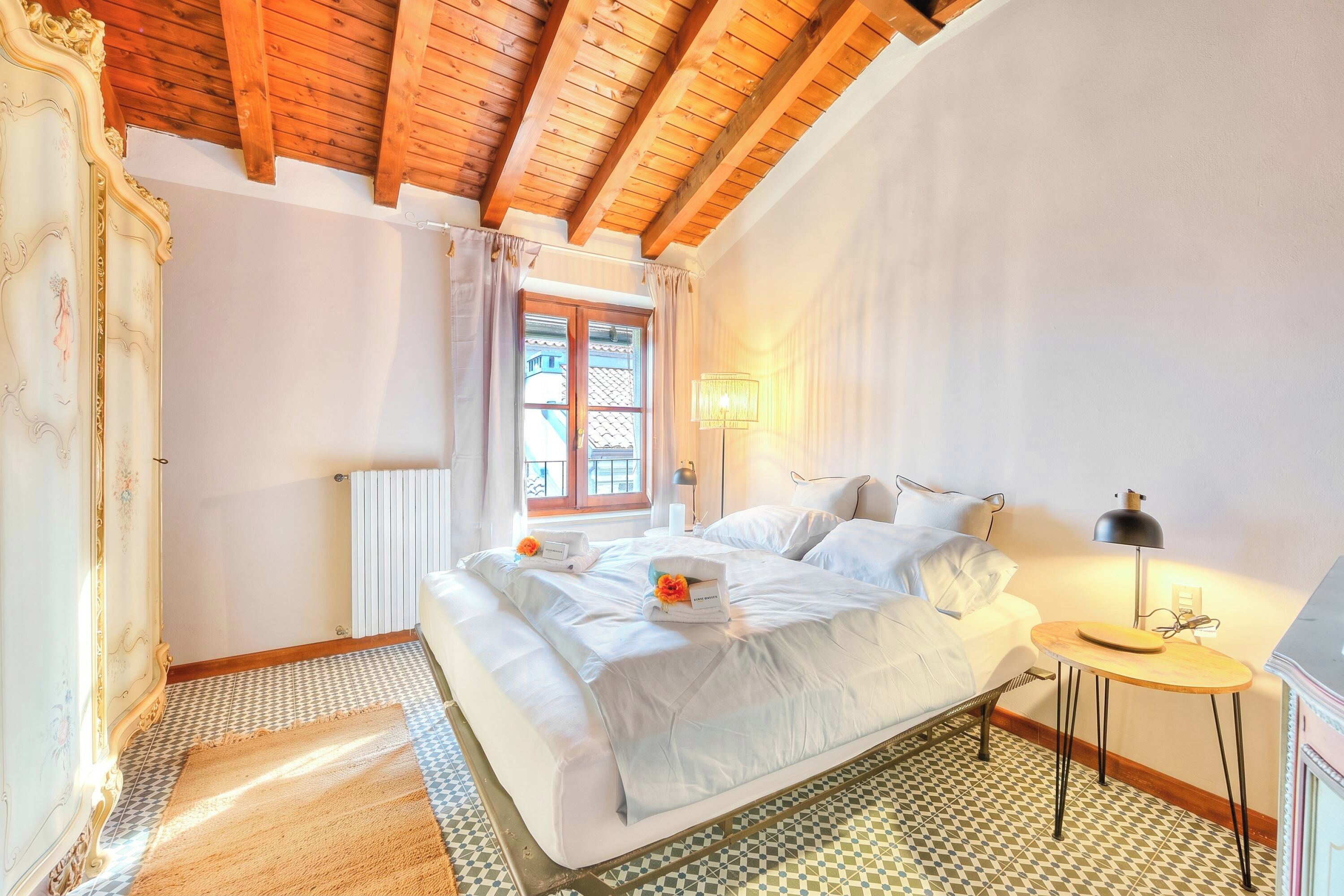 A bedroom with a wooden ceiling, a bed with white linens, a small bedside table, and a radiator.