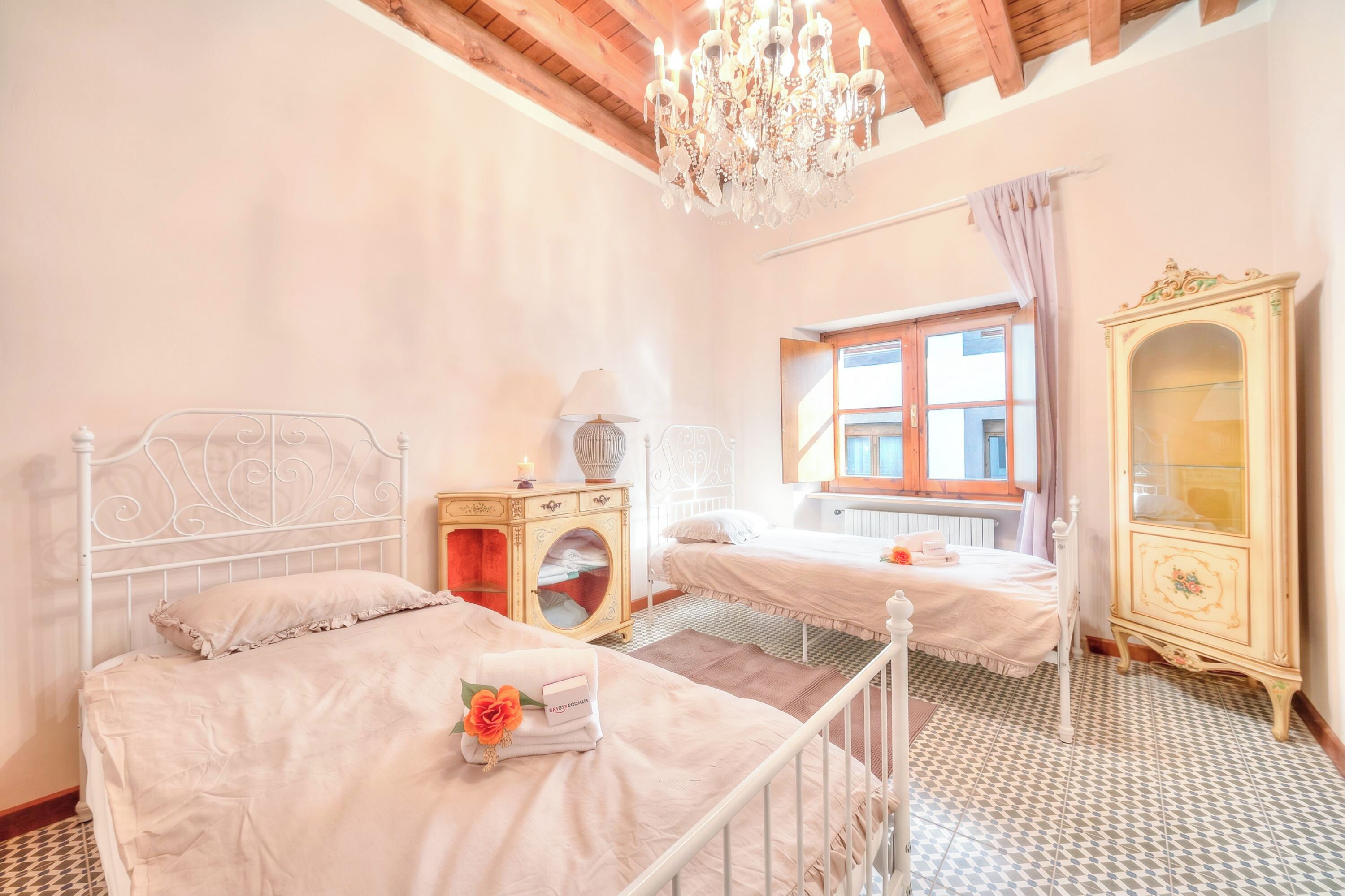 Une chambre avec deux lits, un lustre, une fenêtre avec des rideaux et une commode ancienne.