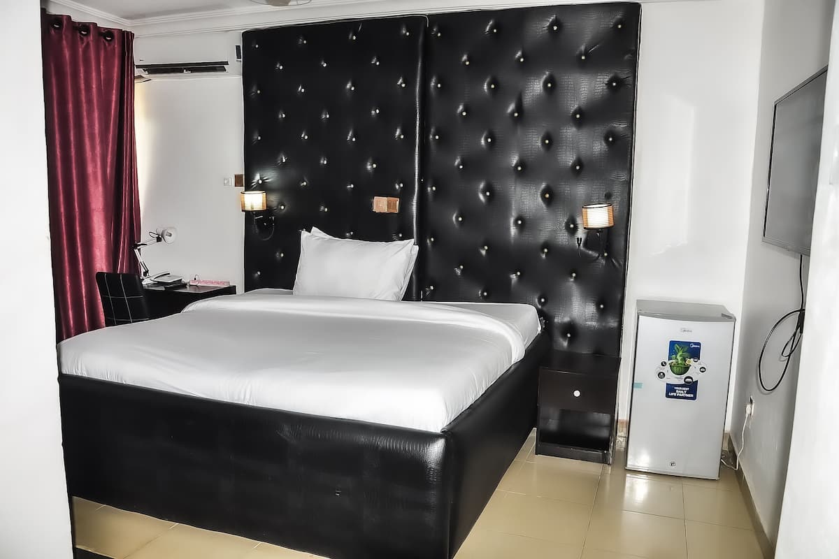 Une chambre d’hôtel avec une tête de lit noire recouverte de tissu, un couvre-lit blanc, une petite table de chevet avec une lampe et un mini-bar.