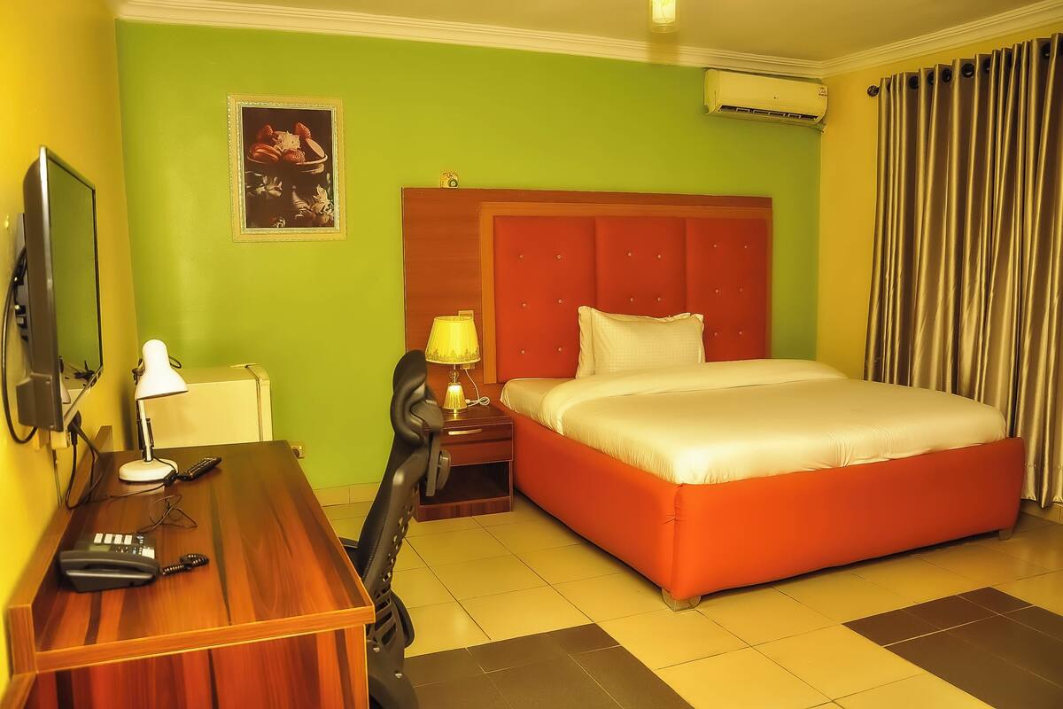 Une chambre d’hôtel avec un bureau en bois, un lit avec une tête de lit rouge, une table de chevet avec une lampe et un climatiseur fixé au mur.