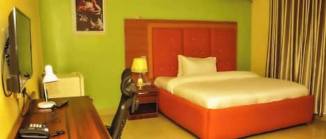 Una habitación de hotel con un escritorio de madera, una cama con cabecera roja, una mesita de noche con lámpara y un aire acondicionado de pared.