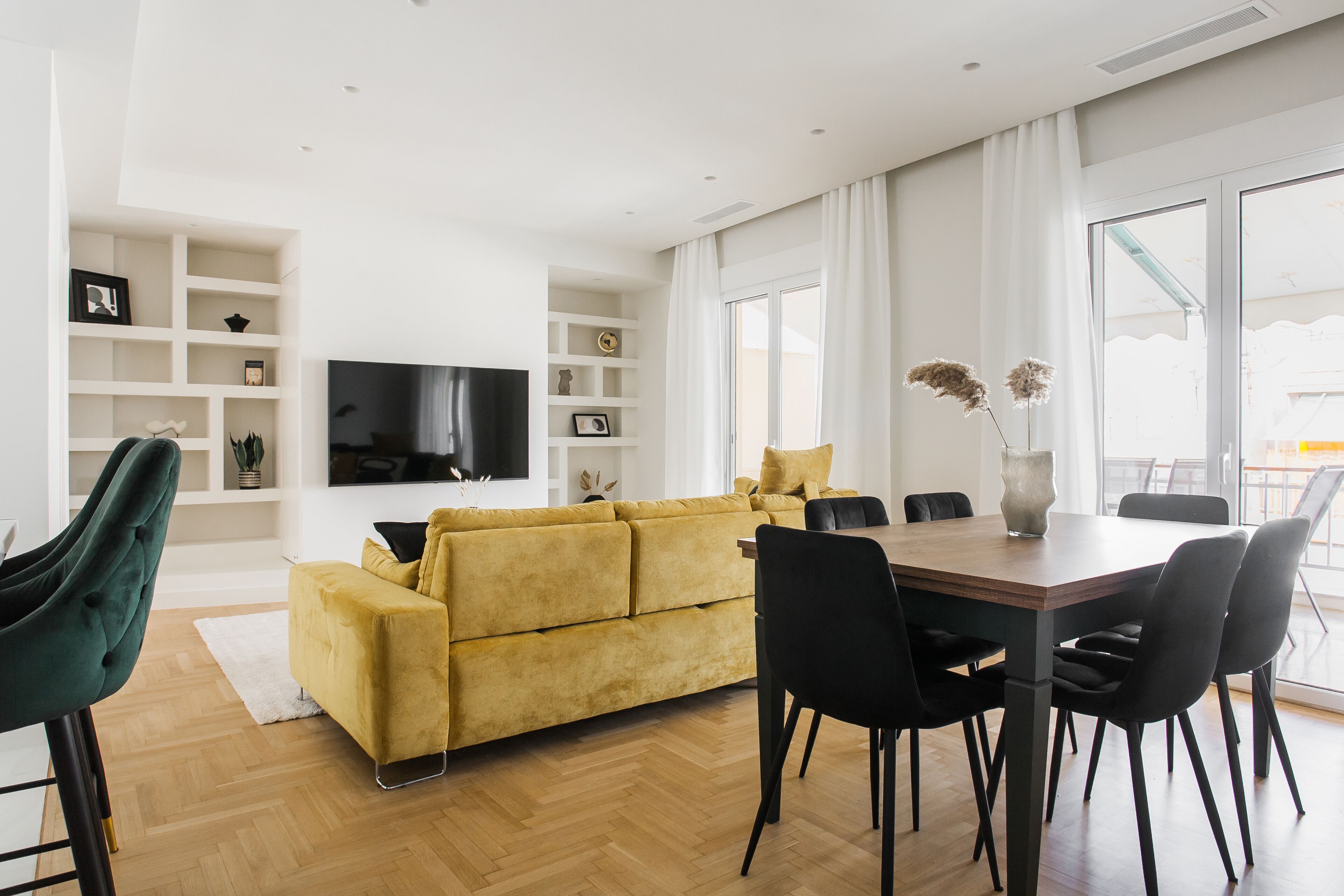 A modern living room with a yellow sofa, a wooden dining table, and black chairs. There is a flat-screen TV mounted on the wall, and a large window with curtains.
