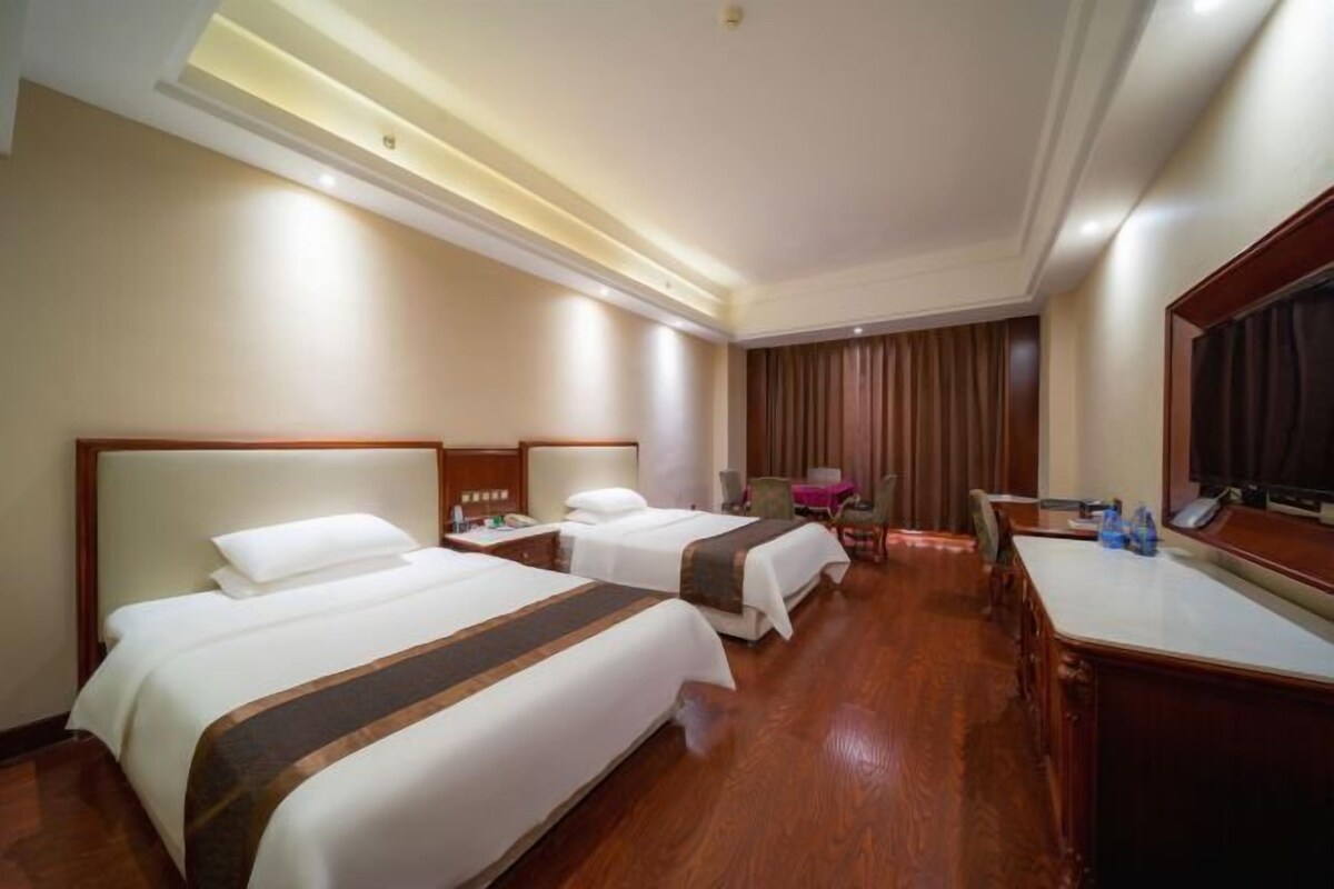 A hotel room with two beds, a desk, a chair, a TV, and a large window with curtains.