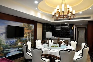 Un coin repas avec une table ronde dressée pour six personnes, entourée de chaises blanches à bordures noires. Un grand tableau représentant un paysage orne le mur, et un lustre est suspendu au plafond.
