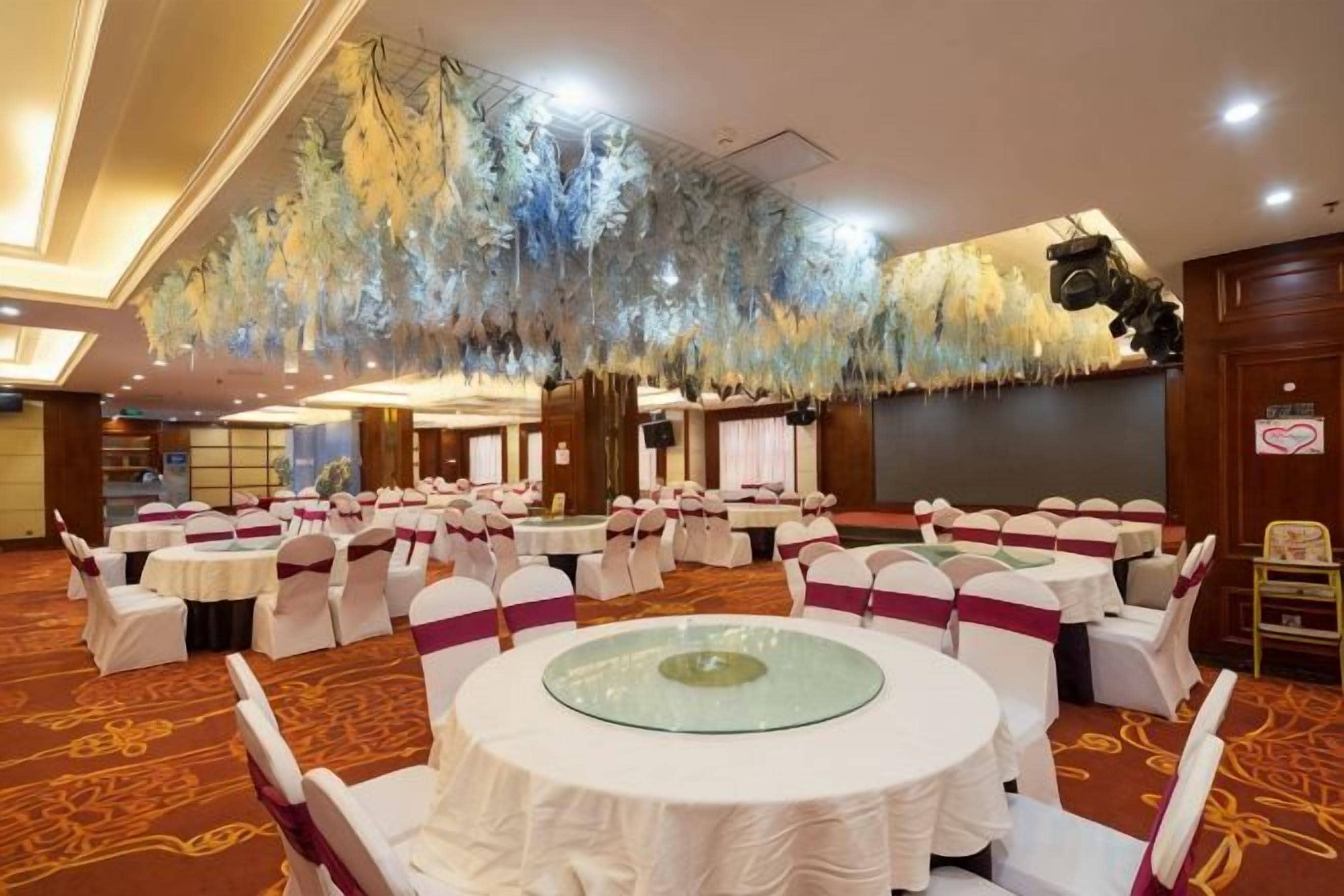 Une salle de banquet avec des tables rondes, des chaises blanches et un plafond orné de décorations suspendues.