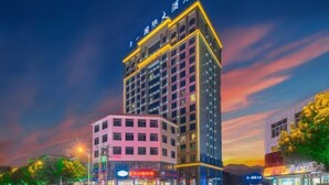 Un hotel de varios pisos con un letrero llamativo en la parte superior, ubicado en una esquina con tiendas contiguas, y vista a la calle con automóviles estacionados.