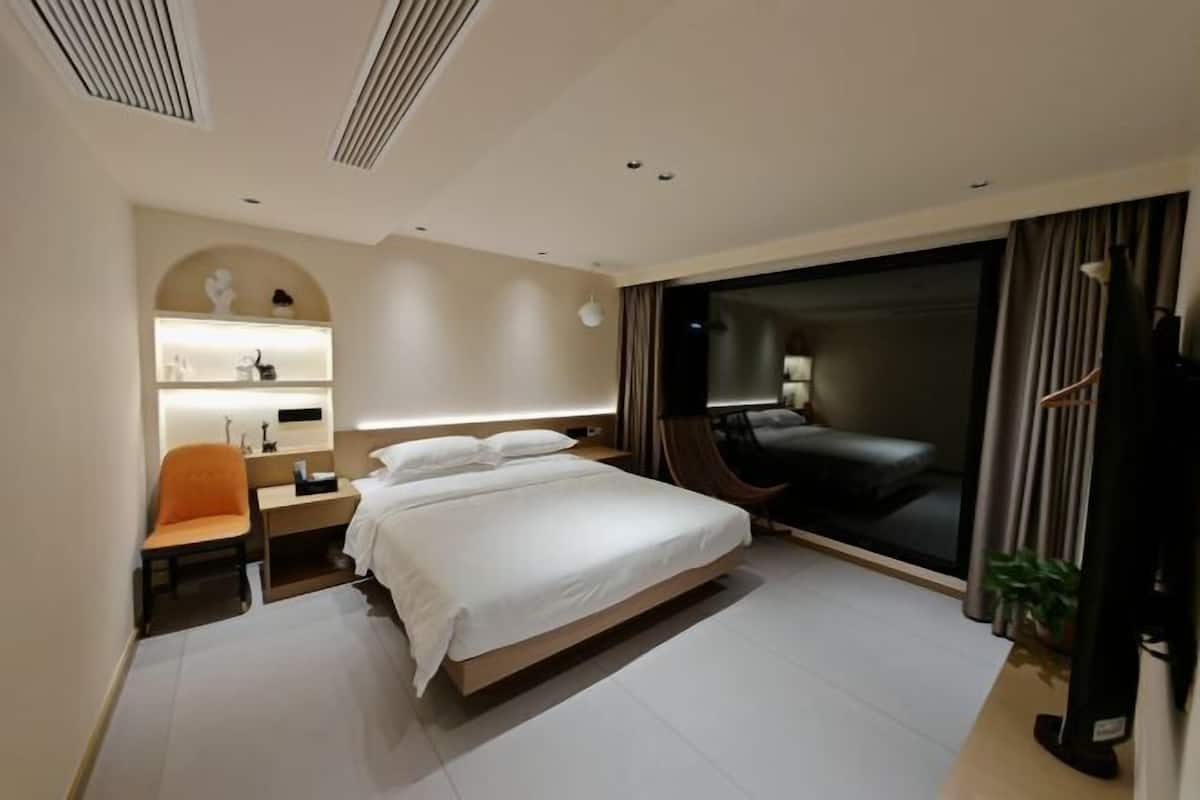 A modern hotel room with a large bed, a desk with a chair, a TV, and a sliding glass door leading to a balcony.