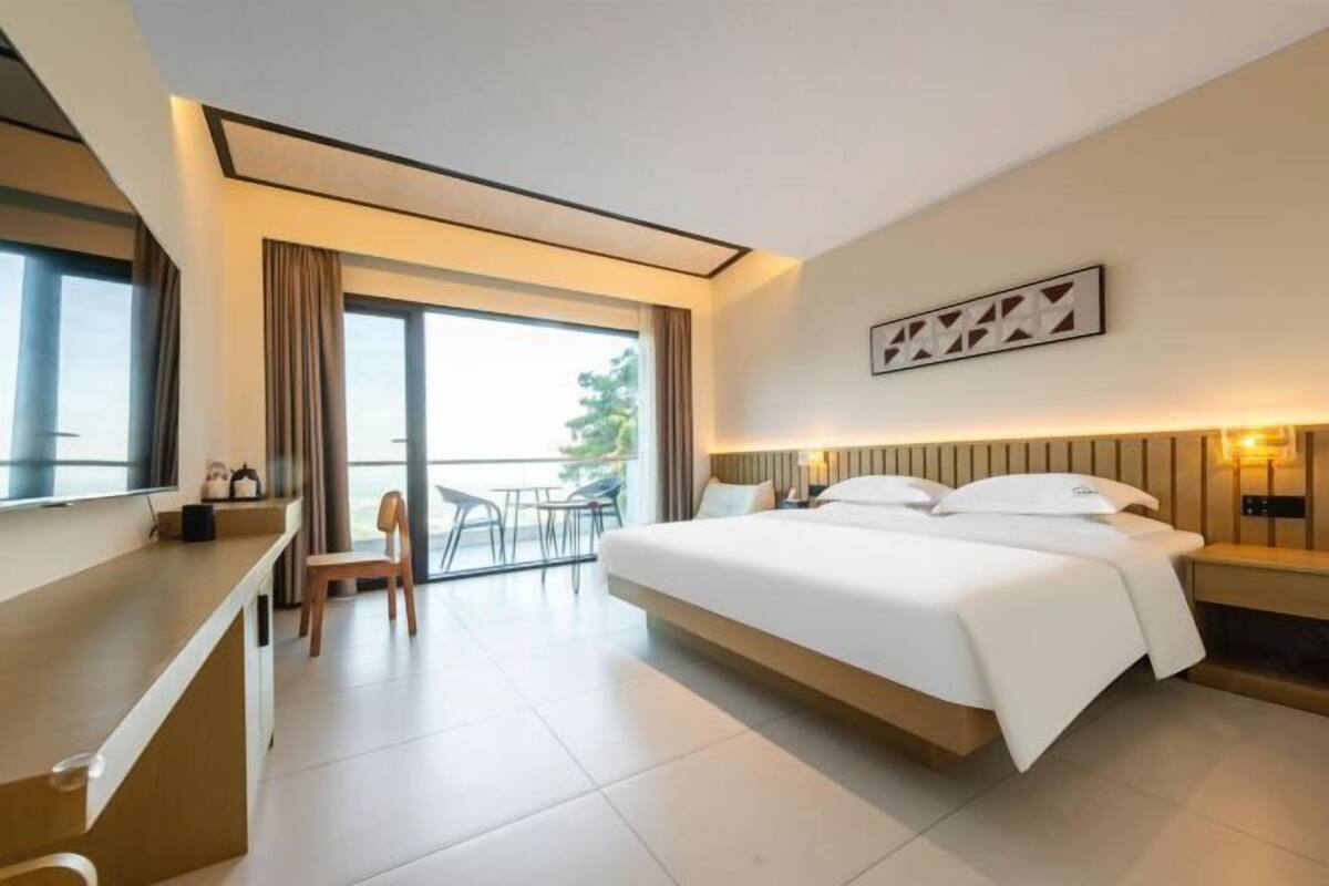 A modern hotel room with a large bed, a desk, and a balcony with a view.