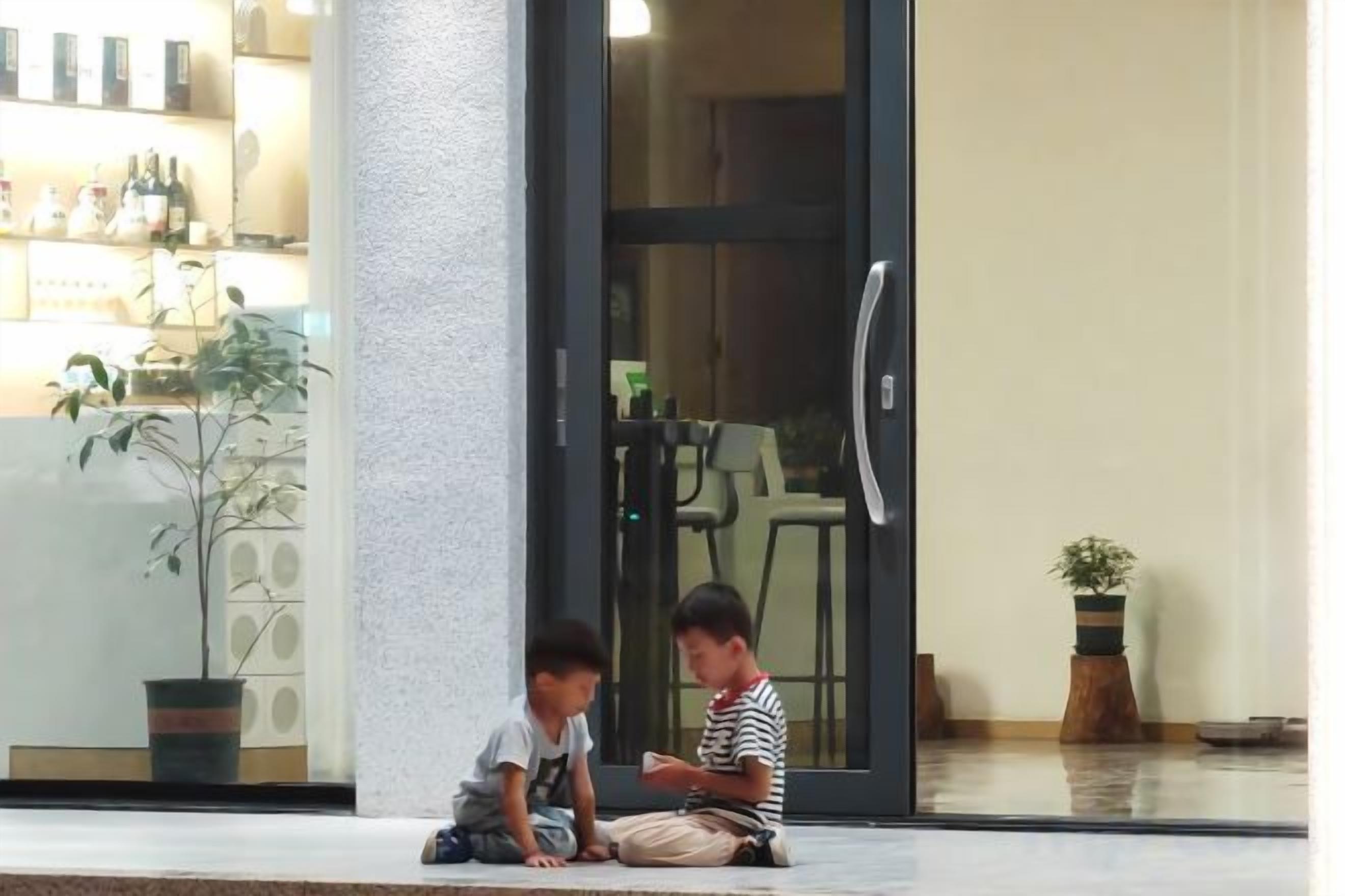Two individuals seated on a ledge outside a building with a glass door and a potted plant nearby.