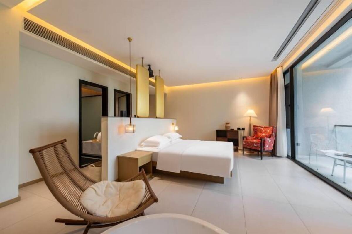 A modern hotel room with a bed, a sitting chair, a small table, a large window, and a view of the outside.