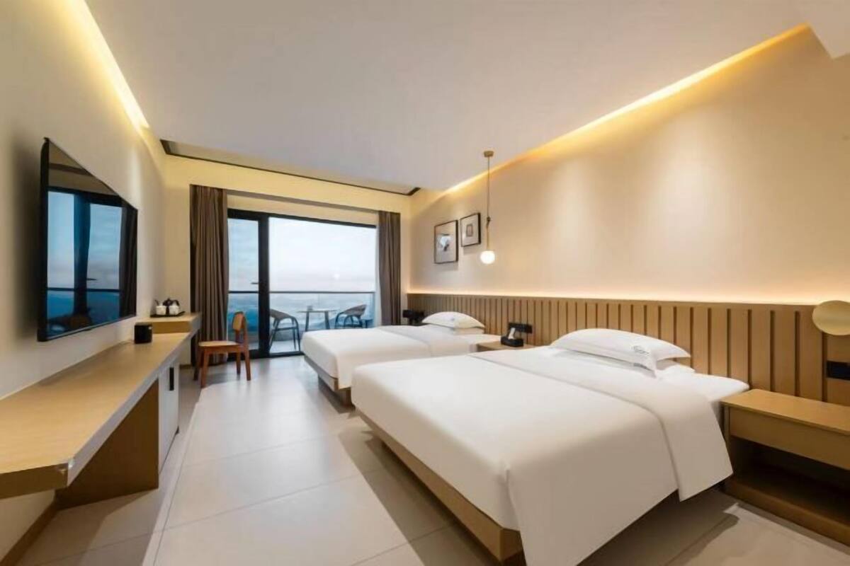 A hotel room with two beds, a desk, a chair, a TV, and a view of the sea.