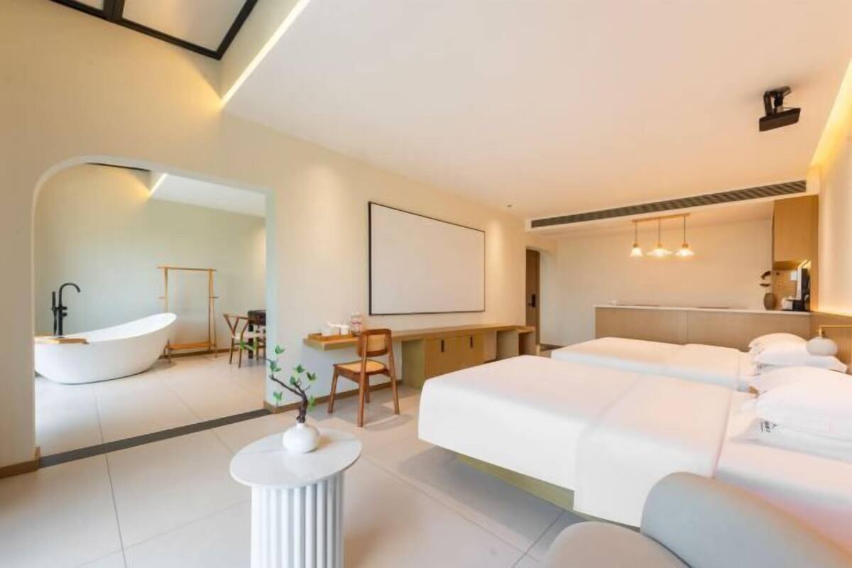 A modern hotel room with a large bed, a sitting area with a desk and chair, a bathtub, and a large mirror.