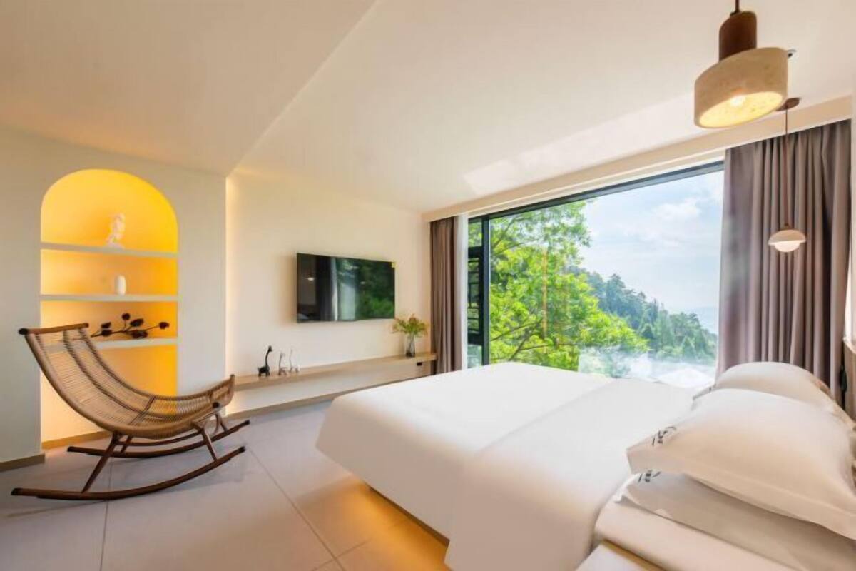 A modern hotel room with a large bed, a rocking chair, a TV, and a view of greenery.