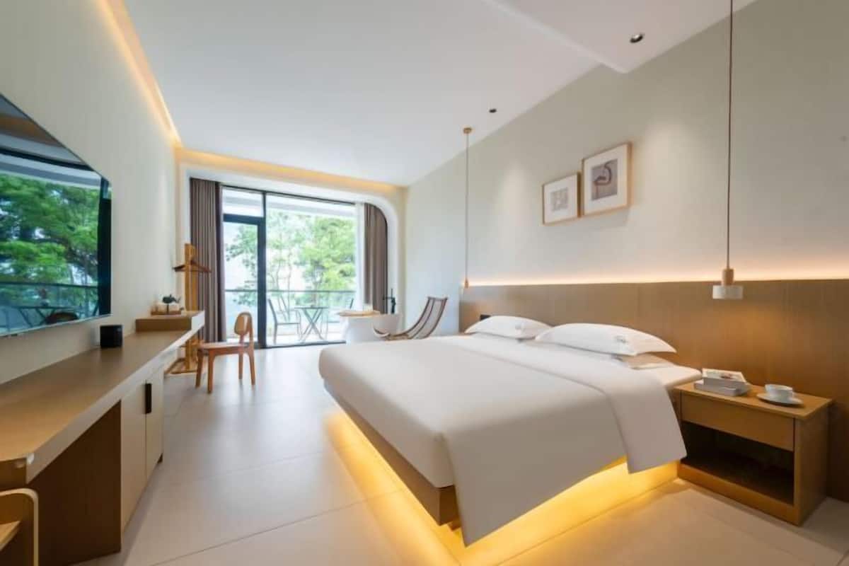A modern hotel room with a bed, bedside table, desk, and a view of the outdoors.