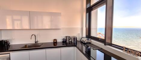A modern kitchen with a stainless steel sink, white cabinets, and a window overlooking the sea.