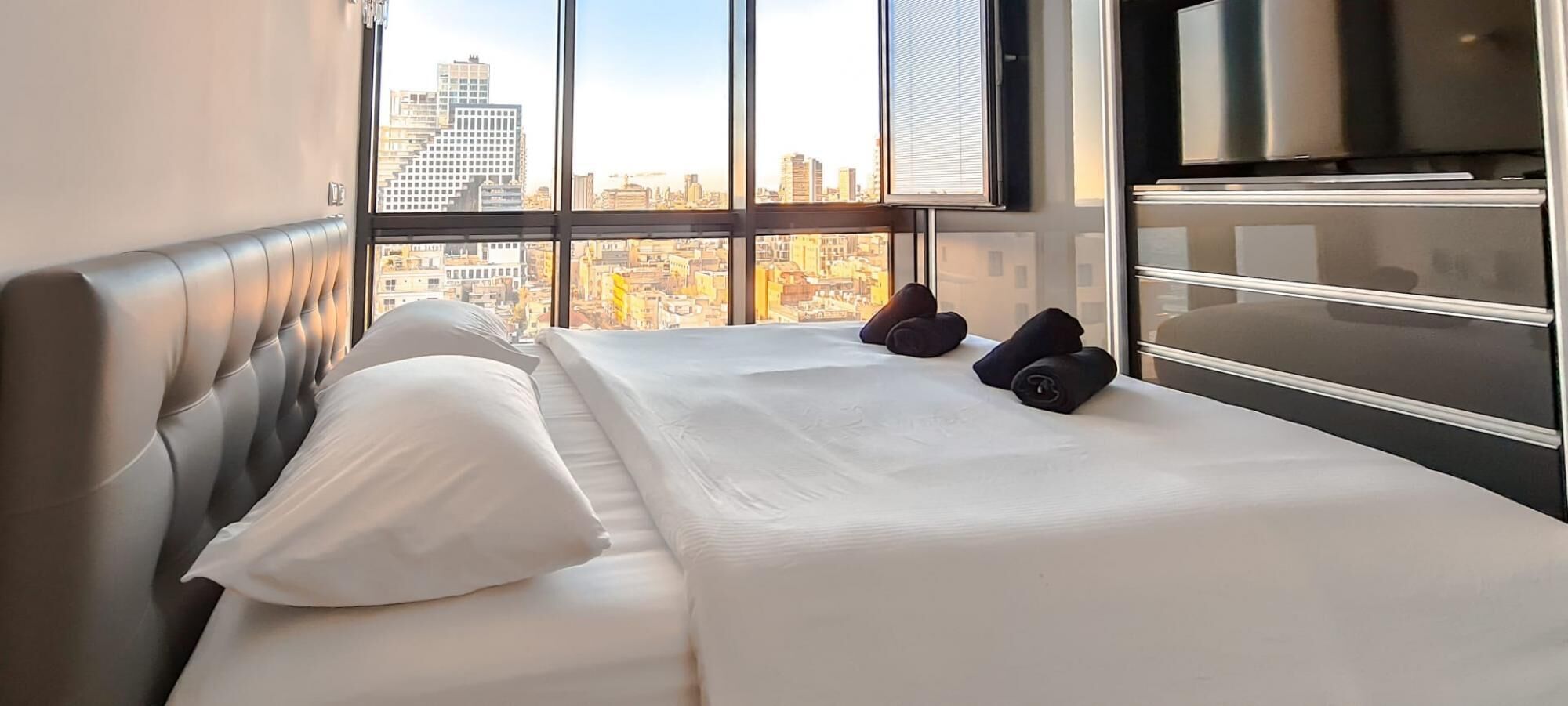 A hotel room with a large bed, a view of the cityscape through floor-to-ceiling windows, a modern headboard, and a minibar.
