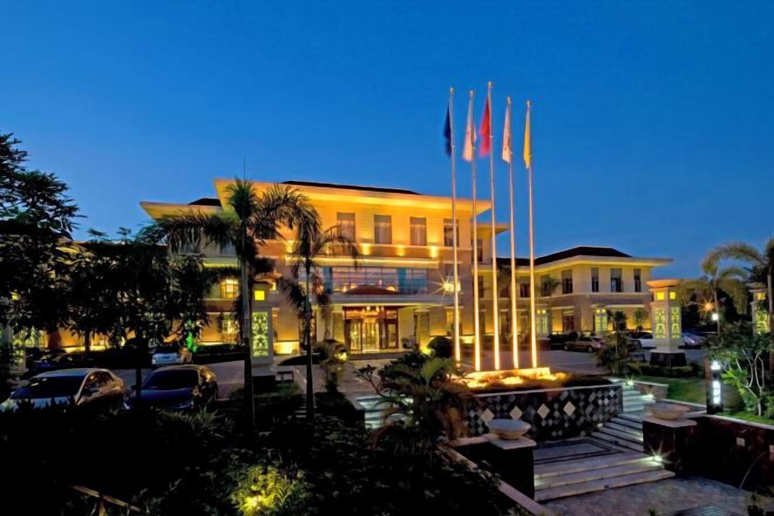 A hotel with a well-lit entrance, a flagpole with flags, and a landscaped garden area.