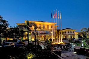 Un hotel con una entrada bien iluminada, un asta con banderas y un jardín ajardinado.