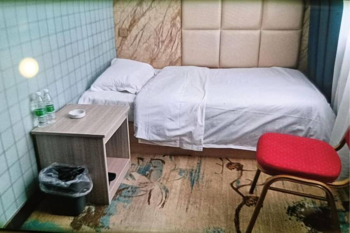 A small, neatly arranged hotel room with a single bed, a small table with a chair, a trash bin, and a wall-mounted light.