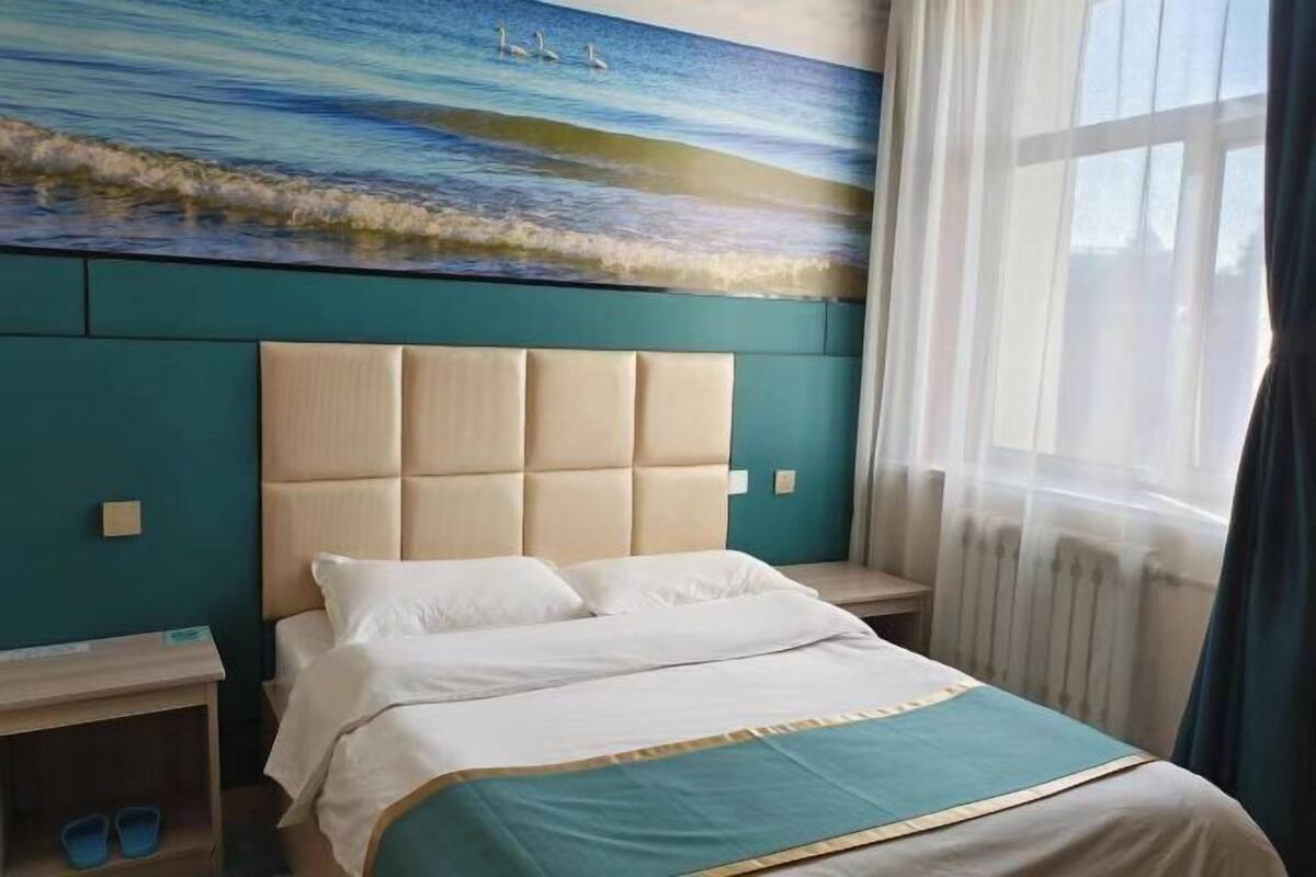 A hotel room with a large bed, a headboard, a nightstand, a window with curtains, and a painting of a beach scene.