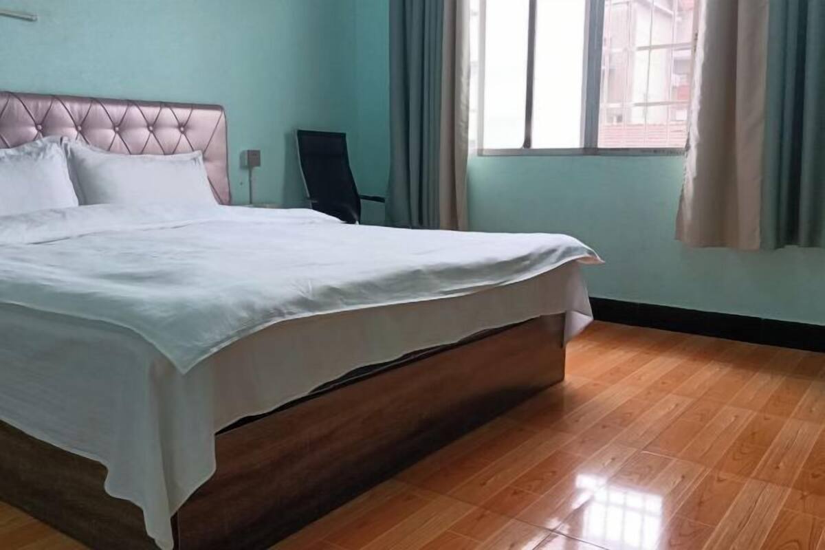 A bedroom with a bed, a chair, a window with curtains, and a wooden floor.