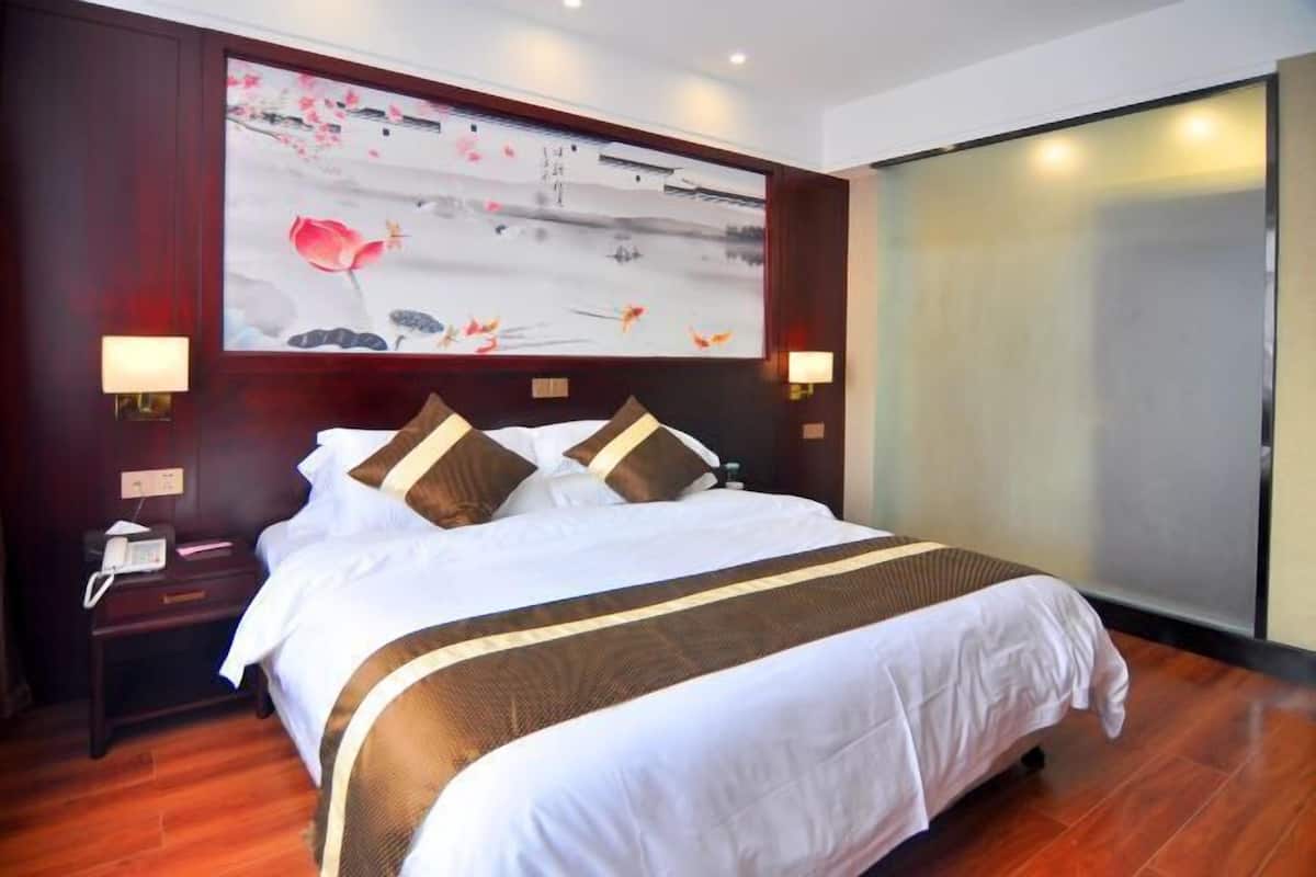 A hotel room with a large bed, wooden headboard, and a painting above it. There is a nightstand with a phone and a lamp.