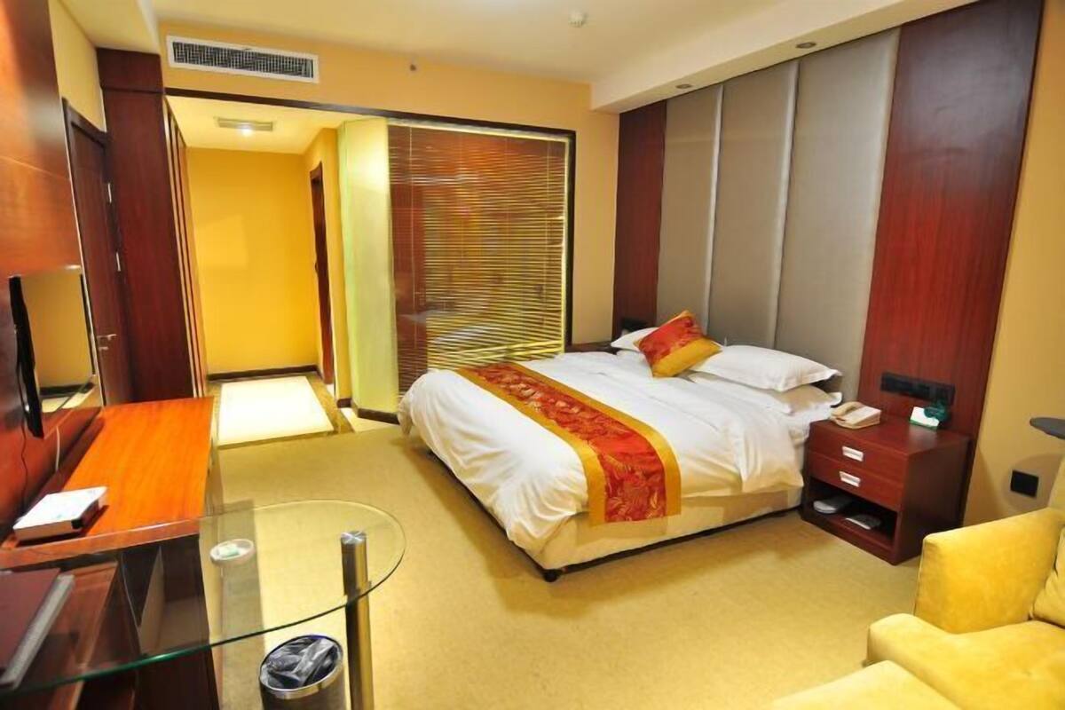 A hotel room with a bed, bedside table, a desk with a chair, a wardrobe, and a glass partition.