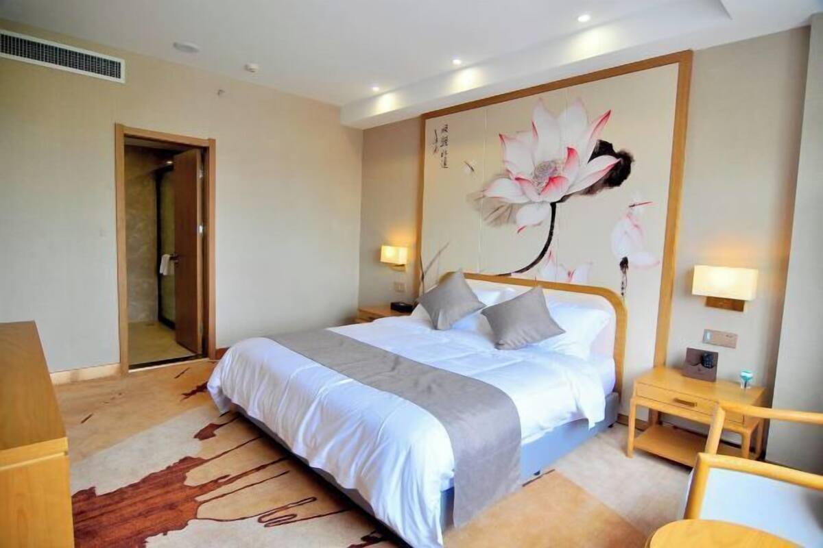 A hotel room with a large bed, bedside tables, a wooden nightstand, a desk, and a wall with a floral painting.