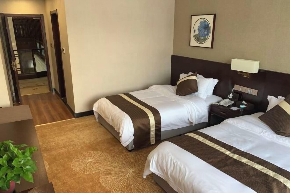 A hotel room with two beds, a nightstand, a lamp, a painting on the wall, and a potted plant.